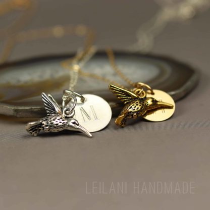 Two hummingbird keepsake necklace, with hummingbird charms, one silver with an "M" engraved charm and one gold with an "H" engraved charm, rest on a smooth stone. Both are on delicate chains. The background is blurred, and "LEILANI HANDMADE" is faintly visible at the bottom.