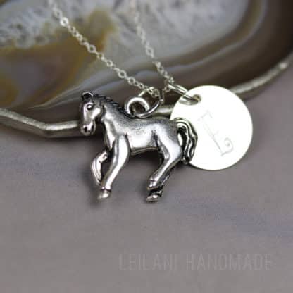 A silver horse keepsake necklace with a detailed horse charm, a polished chain and a round disc engraved with the letter “E.” The necklace rests on a stone dish with an agate pattern. The words "LEILANI HANDMADE" are faintly visible in the lower right corner.