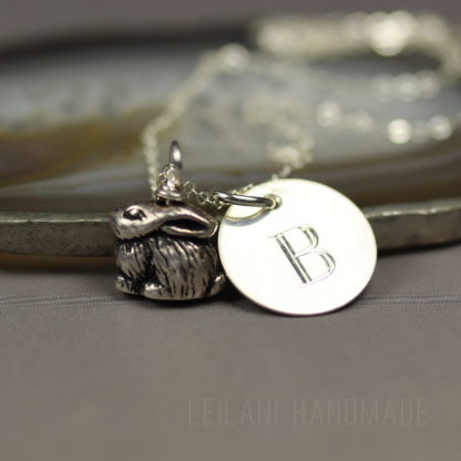 A silver personalized bunny charm necklace with a small, detailed rabbit charm and a round pendant engraved with the letter "B," resting on a smooth stone surface. The background is soft and neutral, emphasizing the jewelry.