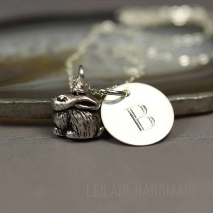 A silver personalized bunny charm necklace with a small, detailed rabbit charm and a round pendant engraved with the letter "B," resting on a smooth stone surface. The background is soft and neutral, emphasizing the jewelry.