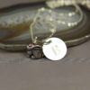 A silver necklace with a small rabbit charm and a round disc engraved with the letter “B,” displayed on a polished agate slice. The background is a soft gray, and “LEILANI HANDMADE” is faintly visible at the bottom.