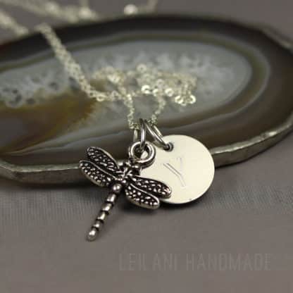 A silver necklace with a round pendant engraved with the letter "V" and a detailed dragonfly charm rests on a polished agate slice. The background is a neutral gray, and the chain is loosely coiled behind the pendants. "LEILANI HANDMADE" is faintly visible.