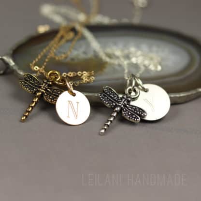 Two dragonfly necklaces rest on a round agate tray. Each necklace has a dragonfly charm and a metal disc featuring an engraved letter (“N” on gold, “Y” on silver). The background is taupe, and “LEILANI HANDMADE” is faintly visible at the bottom.