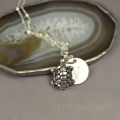 A silver turtle pendant and a round charm engraved with the letter "V" rest on a silver chain, displayed on a sliced agate stone with earthy tones. The background is light gray, and "LEILANI HANDMADE" is subtly visible at the bottom.