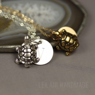 Custom turtle necklace: Two turtle-shaped pendants, one silver and one gold, lie on chains beside a round, engraved disc on a gray stone surface. The background features a sliced agate. The words "LEILANI HANDMADE" are faintly visible in the lower right corner.
