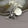 A silver frog charm necklace with a round disc engraved with the letter "G" rests on a polished agate slice; the background is neutral, and "Leilani Handmade" is faintly visible at the bottom.