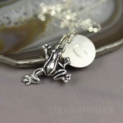 A silver frog charm necklace with a round disc engraved with the letter "G" rests on a polished agate slice; the background is neutral, and "Leilani Handmade" is faintly visible at the bottom.
