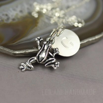 A silver frog charm necklace with a round disc engraved with the letter "G" rests on a polished agate slice; the background is neutral, and "Leilani Handmade" is faintly visible at the bottom.
