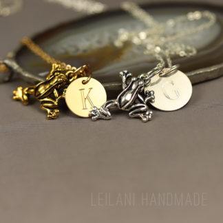 Two leaping frog keepsake necklaces with frog-shaped pendants and round initial charms—one gold with a "K," and one silver with a "G." The necklaces rest on a smooth stone slice against a neutral background. "LEILANI HANDMADE" is faintly visible at the bottom.