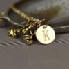 A gold frog charm and a round gold pendant engraved with the letter “K” hang together on a delicate chain. The necklace is displayed on a gray surface, with a blurred agate stone in the background. The text “LEILANI HANDMADE” appears faintly at the bottom.