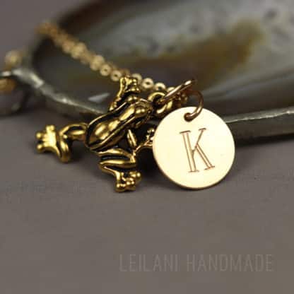A gold frog charm and a round gold pendant engraved with the letter “K” hang together on a delicate chain. The necklace is displayed on a gray surface, with a blurred agate stone in the background. The text “LEILANI HANDMADE” appears faintly at the bottom.