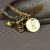 A gold frog charm and a round gold pendant engraved with the letter “K” hang together on a delicate chain. The necklace is displayed on a gray surface, with a blurred agate stone in the background. The text “LEILANI HANDMADE” appears faintly at the bottom.