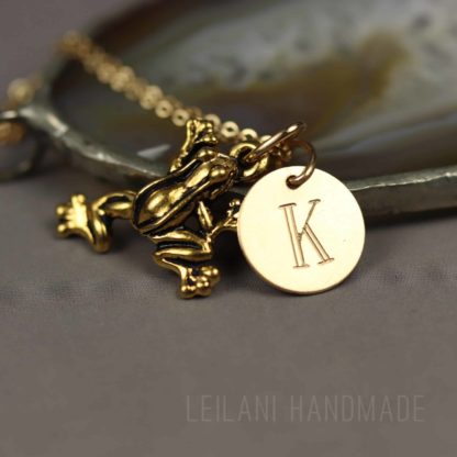 A gold frog charm and a round gold pendant engraved with the letter “K” hang together on a delicate chain. The necklace is displayed on a gray surface, with a blurred agate stone in the background. The text “LEILANI HANDMADE” appears faintly at the bottom.