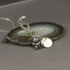 A silver teddy bear charm necklace with a round engraved pendant rests on a polished agate slice. The background is a soft, matte gray. The necklace chain is delicate, and the pendant features the engraved letter “B.” The words "LEILANI HANDMADE" appear faintly at the bottom.
