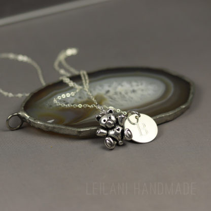A silver teddy bear charm necklace with a round engraved pendant rests on a polished agate slice. The background is a soft, matte gray. The necklace chain is delicate, and the pendant features the engraved letter “B.” The words "LEILANI HANDMADE" appear faintly at the bottom.