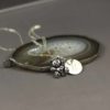 A silver teddy bear charm necklace with a round engraved pendant rests on a polished agate slice. The background is a soft, matte gray. The necklace chain is delicate, and the pendant features the engraved letter “B.” The words "LEILANI HANDMADE" appear faintly at the bottom.