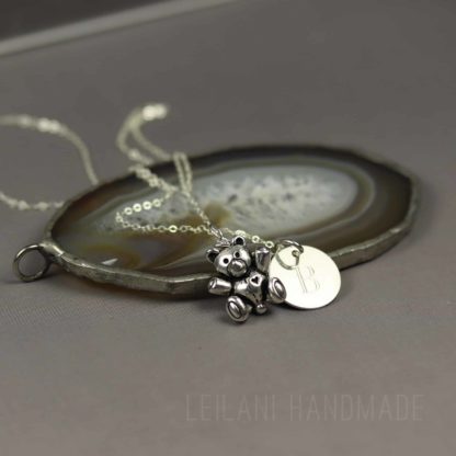 A silver teddy bear charm necklace with a round engraved pendant rests on a polished agate slice. The background is a soft, matte gray. The necklace chain is delicate, and the pendant features the engraved letter “B.” The words "LEILANI HANDMADE" appear faintly at the bottom.