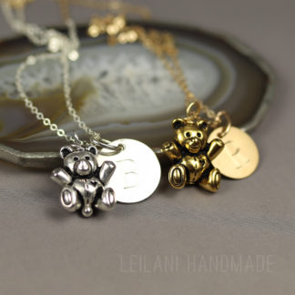 Two necklaces rest on a gray surface: one is silver with a teddy bear charm and round disc engraved with "B," and the other is gold with a similar teddy bear and disc engraved with "L." Both lie in front of a polished stone slice. Text reads "LEILANI HANDMADE.