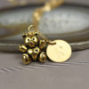 A gold teddy bear charm with a heart on its chest sits beside a round gold pendant engraved with the letter "R," both hanging from a delicate chain. The background is softly blurred, and “LEILANI HANDMADE” is faintly visible at the bottom.