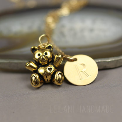 A gold teddy bear charm with a heart on its chest sits beside a round gold pendant engraved with the letter "R," both hanging from a delicate chain. The background is softly blurred, and “LEILANI HANDMADE” is faintly visible at the bottom.