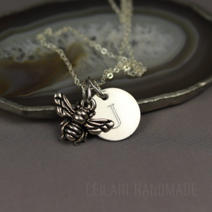 A silver necklace with two pendants: a detailed bee charm and a round disc engraved with the letter "J." The necklace is displayed on a slice of agate and rests on a gray surface. The words "LEILANI HANDMADE" are faintly visible at the bottom.
