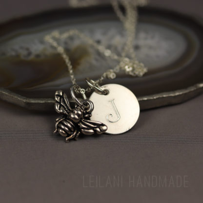 A silver necklace with a bee charm and a round pendant engraved with the letter “J” rests on a polished agate slice. The background is gray, and “LEILANI HANDMADE” is faintly visible at the bottom.