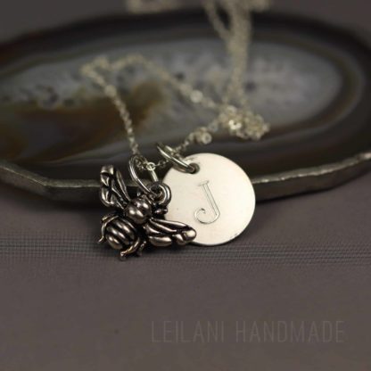 A silver necklace with a bee charm and a round pendant engraved with the letter “J” rests on a polished agate slice. The background is gray, and “LEILANI HANDMADE” is faintly visible at the bottom.
