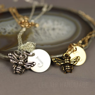 Two bee keepsake necklaces with bee charms and round initial pendants rest on a polished agate slice. One necklace is silver with a silver “J” pendant, the other is gold with a gold “J” pendant. The background is neutral and text reads “LEILANI HANDMADE.”.