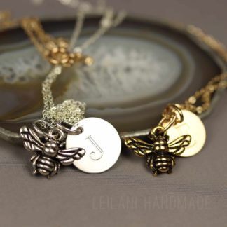 Two bee keepsake necklaces with bee charms and round initial pendants rest on a polished agate slice. One necklace is silver with a silver “J” pendant, the other is gold with a gold “J” pendant. The background is neutral and text reads “LEILANI HANDMADE.”.