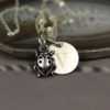 A silver necklace with two charms: a ladybug and a round disc engraved with the letter "V." The jewelry rests on a polished agate slice with blurred text "LEILANI HANDMADE" visible beneath. The photo has a soft, muted background.