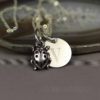 A silver necklace with two charms: a ladybug and a round disc engraved with the letter "V." The jewelry rests on a polished agate slice with blurred text "LEILANI HANDMADE" visible beneath. The photo has a soft, muted background.