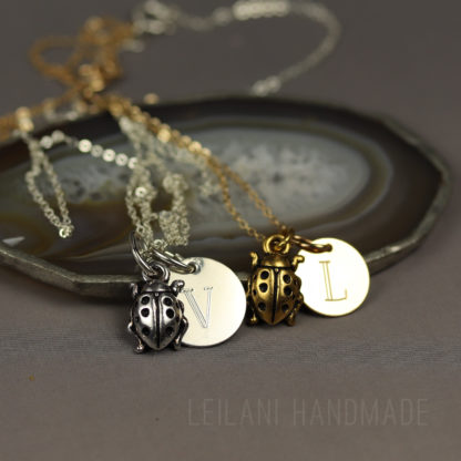 Two necklaces rest on a sliced agate dish: one silver with a round pendant engraved “V” and a silver ladybug charm, and one gold with a round pendant engraved “L” and a gold ladybug charm. “LEILANI HANDMADE” is faintly visible in the background.