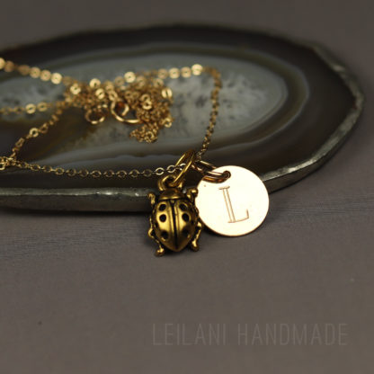 A gold necklace with a ladybug charm and a round pendant engraved with the letter "L" rests on an agate slice. The background is a smooth, dark surface, and the text "LEILANI HANDMADE" is faintly visible at the bottom.