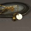 A gold necklace with a ladybug charm and a round pendant engraved with the letter "L" rests on an agate slice. The background is a smooth, dark surface, and the text "LEILANI HANDMADE" is faintly visible at the bottom.