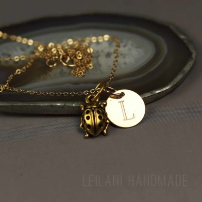 A gold necklace with a ladybug charm and a round pendant engraved with the letter "L" rests on an agate slice. The background is a smooth, dark surface, and the text "LEILANI HANDMADE" is faintly visible at the bottom.