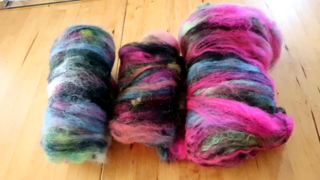 Three rolls of colorful, fluffy carded wool batts rest on a wooden surface. The wool blends vibrant shades of pink, blue, green, black, and purple in a soft, textured swirl, ready for spinning.