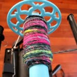 A close-up of a spinning wheel bobbin wrapped with colorful yarn, mostly black with pink, green, and yellow strands. The bobbin is bright blue with a decorative end, and the spinning wheel sits on a wooden floor.