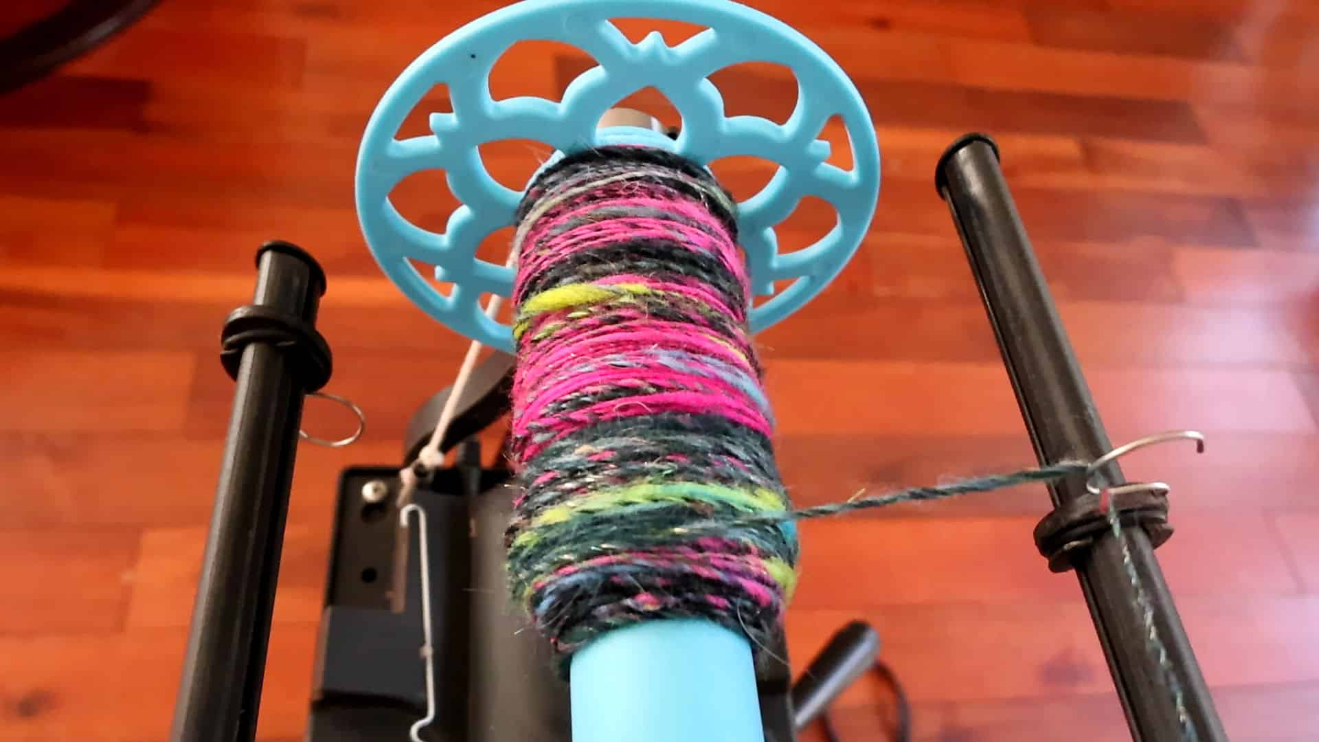 A close-up of a spinning wheel bobbin wrapped with colorful yarn, mostly black with pink, green, and yellow strands. The bobbin is bright blue with a decorative end, and the spinning wheel sits on a wooden floor.