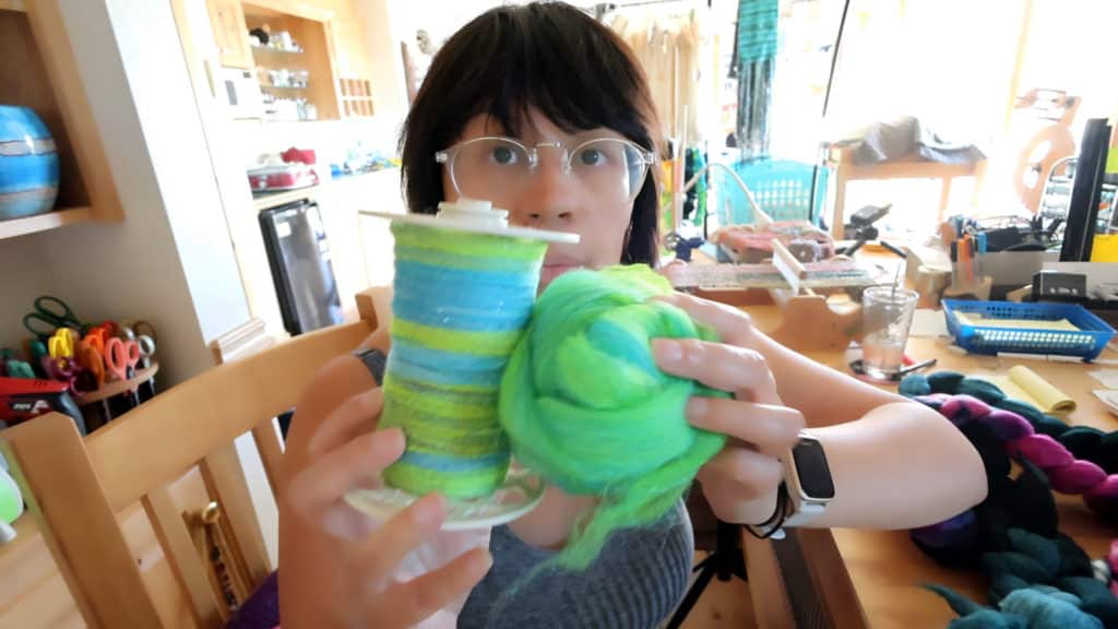 A person with glasses and dark hair holds up a spool of blue and green variegated yarn in one hand and a matching ball of fiber in the other. They sit at a table in a brightly lit craft room with supplies and colorful materials around them. The photo was taken in part to compare two different blends of wool fiber on a hackle for spinning inspiration.
