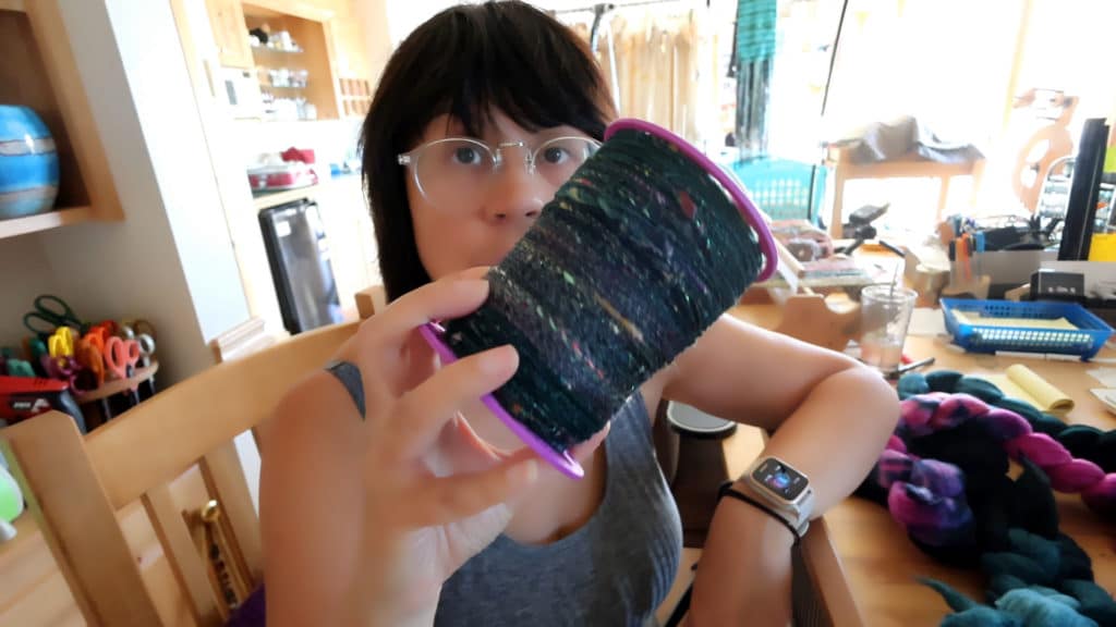 A person with glasses and dark hair holds up a large spool of multicolored yarn in a well-lit, cozy craft room filled with supplies and tools. They wear a smartwatch and a sleeveless gray top, focusing on the yarn.