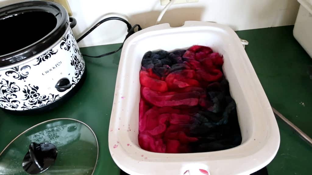 The "ugly dye" has been added to the roving and is now ready to set. A white roasting pan filled with wool roving being dyed in shades of blue and red sits on a green counter. Next to it is a patterned black-and-white Crock-Pot with its glass lid resting nearby. Electrical cords and outlets are visible in the background.