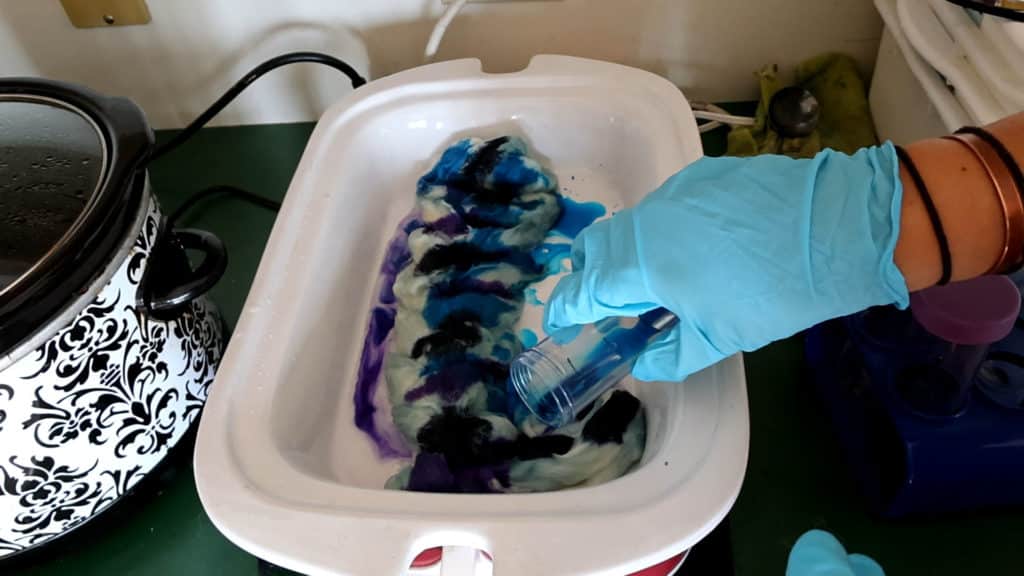 A hand wearing a blue glove pours blue liquid dye onto white wool in a white dish, dyeing roving for spinning. The wool is also streaked with black and purple dye. A patterned slow cooker is visible on the left, and various containers and tools are on the green countertop.