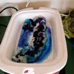 A white tray holds wet wool dyed with blue, purple, and black colors, creating swirling patterns. The tray is on a green counter near a black-and-white slow cooker, a green cloth, utensils, and colorful containers.