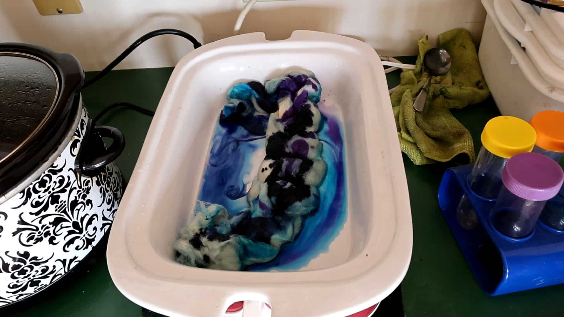 A white tray holds wet wool dyed with blue, purple, and black colors, creating swirling patterns. The tray is on a green counter near a black-and-white slow cooker, a green cloth, utensils, and colorful containers.