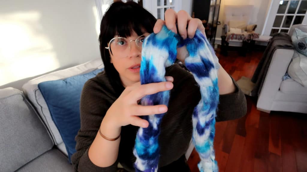 A person with dark hair and glasses sits on a gray couch, holding up the not so ugly dyed roving for spinning. The roving colors are blue, white, and purple with a tie dyed look. The room has wooden floors, a blue cushion, and furniture visible in the softly-lit background.