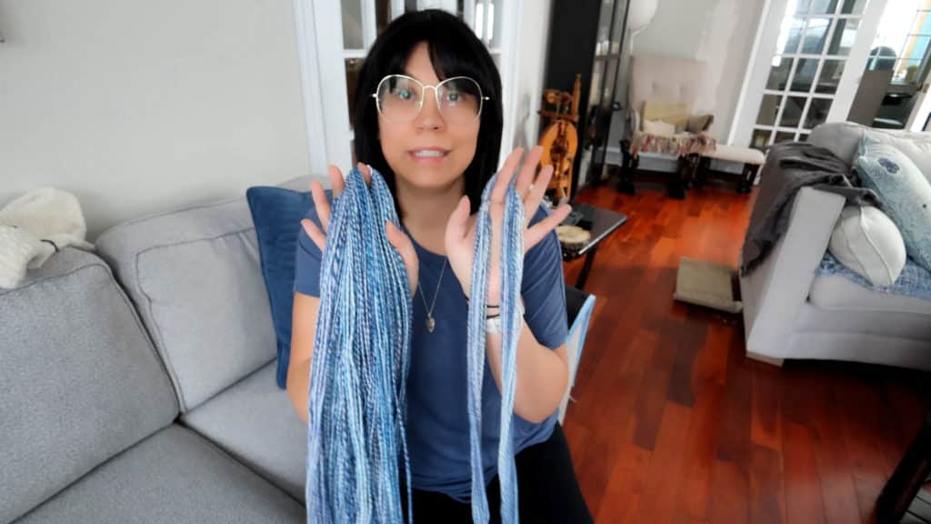 A woman with glasses and dark hair sits on a light gray sofa in a cozy living room, holding up two sections of blue-and-white yarn with both hands. The room has wooden floors, a spinning wheel, and a pile of blankets on the right.