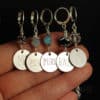 A hand holds five silver stitch markers, each with a round engraved charm and beaded accents. The charms read “M1,” “PSSO,” “PURL,” “KNIT,” and “YO.“ The beads above each charm are various colors and designs. Background is dark.