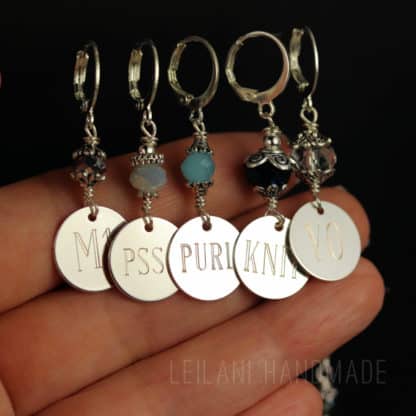 A hand holds five silver stitch markers, each with a round engraved charm and beaded accents. The charms read “M1,” “PSSO,” “PURL,” “KNIT,” and “YO.“ The beads above each charm are various colors and designs. Background is dark.