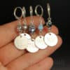 A hand holds four handmade stitch markers with silver clasps, decorative beads, and round metal tags engraved with "SSK," "P2TOG," "M1L," and "M1R." The background is dark, highlighting the intricate details of the markers.