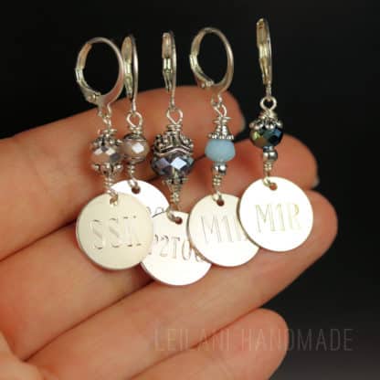 A hand holds four handmade stitch markers with silver clasps, decorative beads, and round metal tags engraved with "SSK," "P2TOG," "M1L," and "M1R." The background is dark, highlighting the intricate details of the markers.